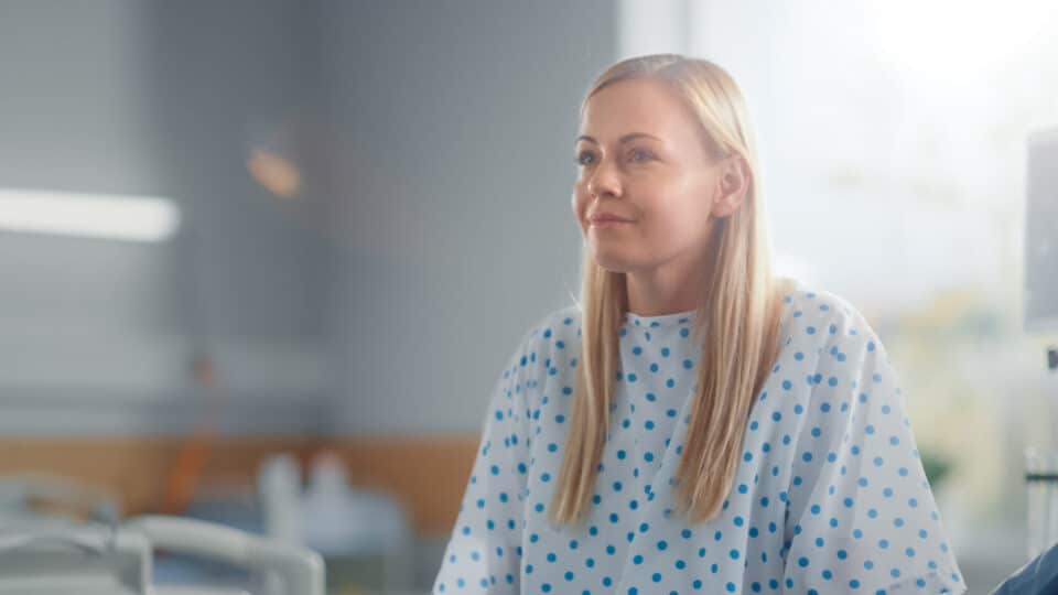 Modern Hospital Ward: Beautiful Caucasian Female Patient Resting On A Bed, Fully Recovering After Successful Surgery, Sickness Or Coronavirus She Smiles. Behind Her Window With A Beautiful Sunny View